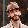 A young guy sporting a neatly trimmed beard, brown fedora hat, and beige jacket stands outside—clearly dressed for the elements. The background’s blurred but you can spot patches of greenery and a bit of blue, like maybe tarps or equipment tucked behind him. - Snow Removal Indianapolis