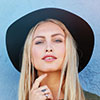Someone with long blonde hair and a wide-brimmed black hat is facing the camera, resting one hand on their chin—kind of like checking the job before plowing starts. They’re standing in front of a textured light blue wall that reminds you of packed snow on a cold morning. - Snow Removal Indianapolis