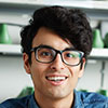 A young individual with dark hair and glasses, smiling as they face the camera, stands in front of a background where green elements are out of focus—much like how we clear away snow to provide a clean view. - Snow Removal Indianapolis